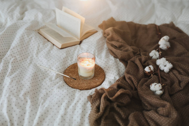 Cozy still life interior details with a book, candle and a cotton twig in warm soft bed. Sweet home - Photo, Image