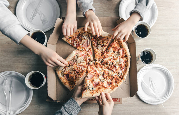 Young people taking slices of hot pizza from cardboard box at table, top view. - Photo, Image