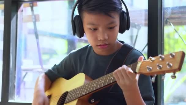 Aasialainen poika oppii soittamaan kitaraa virtuaalisessa kokouksessa pelata musiikkia verkossa yhdessä ystävän tai opettajan kanssa videoneuvottelussa kannettavan tietokoneen kanssa verkossa, Viestintä Internetin kautta Oppiminen käsite - Materiaali, video
