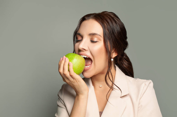 Gri arka planda yeşil bir elmayı ısıran genç kadın. - Fotoğraf, Görsel