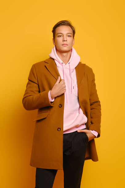 Handsome stylish man in brown coat and pink hoodie posing over yellow studio background - 写真・画像