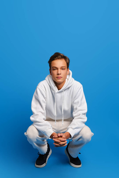Attractive man wearing white sweatshirt and sweatpants is squatting over blue studio background - Photo, Image