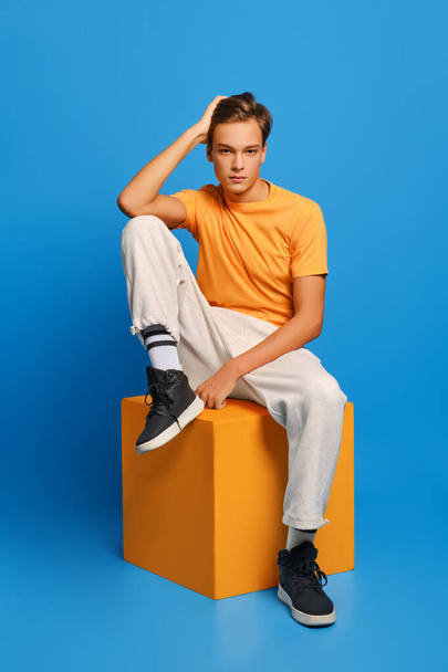 Positive man in orange undershirt and white sweatpants sits on wooden cube over blue studio background - Photo, Image