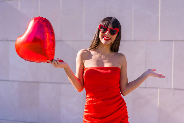 Brunette caucasian girl having fun on Valentine's day with a heart balloon, sunglasses and red dress - Foto, Imagem