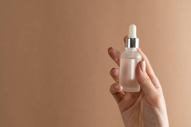 Graceful hand of a girl with thin fingers and a trendy pastel beige nude manicure on a brown background holds a face oil serum - Foto, imagen