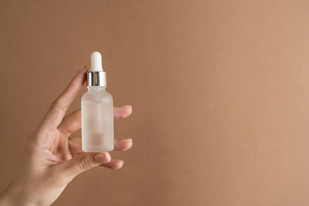 Graceful hand of a girl with thin fingers and a trendy pastel beige nude manicure on a brown background holds a face oil serum - Fotografie, Obrázek