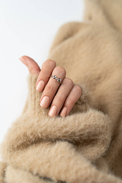 Graceful thin fingers of a girl with a trendy pastel nude manicure and a fluffy soft sweater and a cute silver ring bow - Фото, изображение