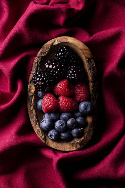 a wooden bowl with blueberries, blackberries and raspberries placed on a red cloth illuminated by a grazing light. View from above - Фото, зображення