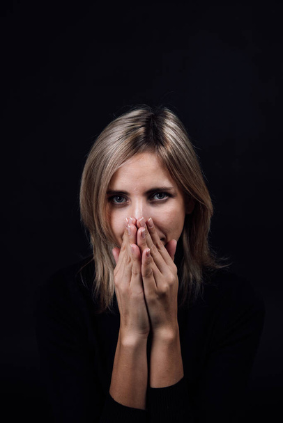 Scared woman with eyes looking at camera, hands hiding nose and mouth in black blouse on black background. Victim of physical and psychological abuse. Gaslighting. Relative aggression - Photo, image