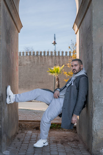 Marocký muž opírající se o zeď ve stylovém šedém kabátě a bílých botách Nike - Fotografie, Obrázek
