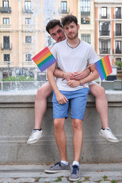 Couple gay étreignant et regardant la caméra tenant des drapeaux LGBT assis par une police. - Photo, image
