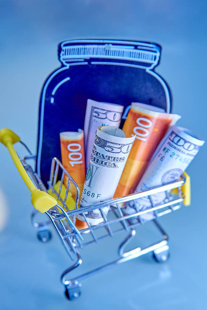 concept - yellow Shopping Cart Trolleys with mock-up of a black jar and money, dollars and Israeli new shekels on light background - Foto, Imagen