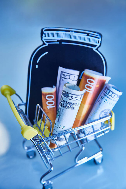 concept - yellow Shopping Cart Trolleys with mock-up of a black jar and money, dollars and Israeli new shekels on light background - Foto, Imagem