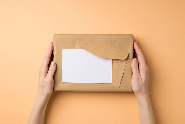 First person top view photo of female hands holding kraft paper giftbox envelope and paper sheet on isolated beige background with blank space - Foto, immagini