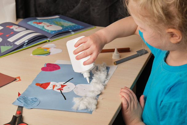 ένα κορίτσι προσχολικής ηλικίας σε ένα μπλε T-shirt κάνει μια απλικέ, ενώ κάθεται στο τραπέζι, ένα χιονάνθρωπο χαρτί τραβηγμένο σε έγχρωμο χαρτί, δημιουργικότητα των παιδιών το χειμώνα, ατοπική δερματίτιδα στα χέρια ενός παιδιού - Φωτογραφία, εικόνα