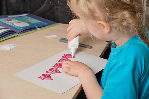 preschooler girl in blue t-shirt makes applique sitting at the table, hearts cut out of colored bougue for card for Valentine's Day, children's creativity, atopic dermatitis on the hands of a child - Fotoğraf, Görsel