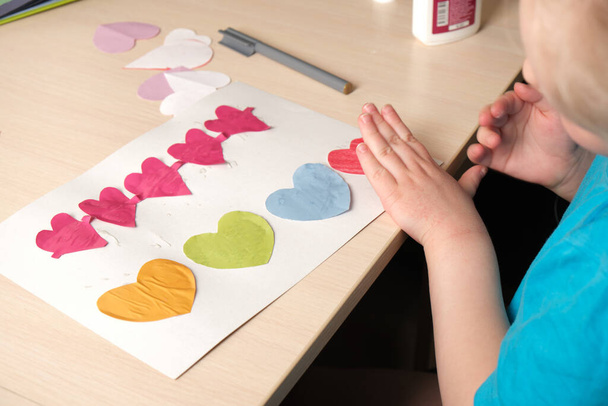 preschooler girl in blue t-shirt makes applique sitting at the table, hearts cut out of colored bougue for card for Valentine's Day, children's creativity, atopic dermatitis on the hands of a child - Foto, afbeelding
