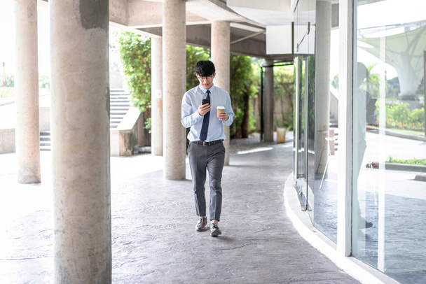 Young business man holding coffee cup and using smartphone to talking business project with partner while walking and looking this mobile to checking e-mail - Foto, afbeelding