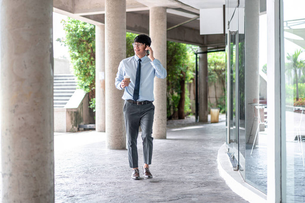 Young business man holding coffee cup and talking on smartphone while walking in office and discussion business project with his partner online - Foto, imagen