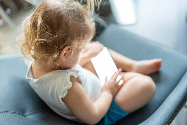 Mosolygó kislány ül a karosszéken, és okos telefont használ. - Fotó, kép