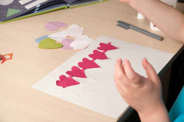 preschooler girl in blue t-shirt makes applique sitting at the table, hearts cut out of colored bougue for card for Valentine's Day, children's creativity, atopic dermatitis on the hands of a child - Foto, Imagen
