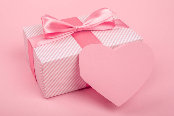 Valentine Day gift in a box wrapped in striped paper and tied with silk ribbon bow and heart shapes greeting card on pink background with copy space for text - Photo, Image