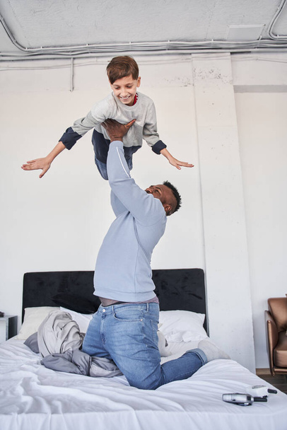 Man holding at the outstretched arms his caucasian son while spending time together at the bedroom - 写真・画像