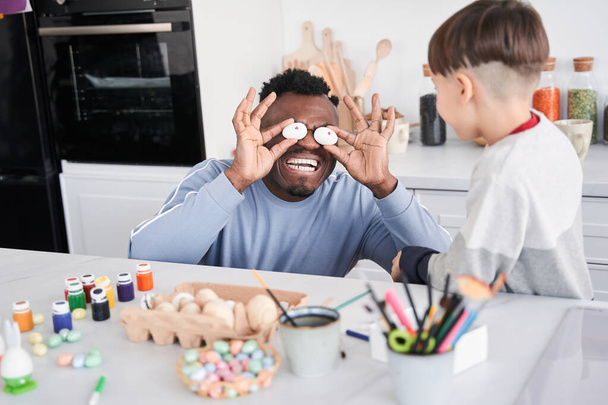Man putting easter eggs instead of his eyes and smiling toothy while joking with his son - Фото, зображення