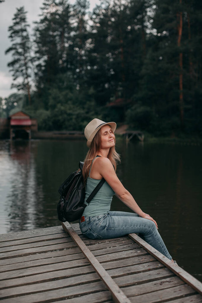 A young blonde girl traveler in jeans and a tank top sits overlooking the lake - Φωτογραφία, εικόνα