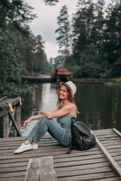 A young blonde girl traveler in jeans and a tank top sits overlooking the lake - Foto, immagini