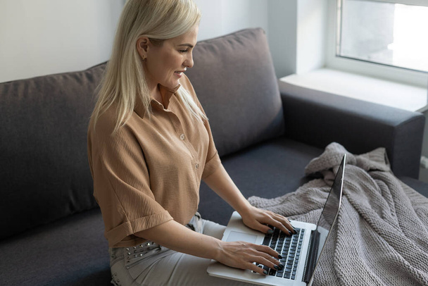 Schöne Blondine arbeitet am Notizbuch und setzt sich zu Hause aufs Sofa - Online-Shops für Cyber-Monday-Verkäufe checken - Technikfrauenkonzept für alternative Büroselbstständigkeit - Foto, Bild