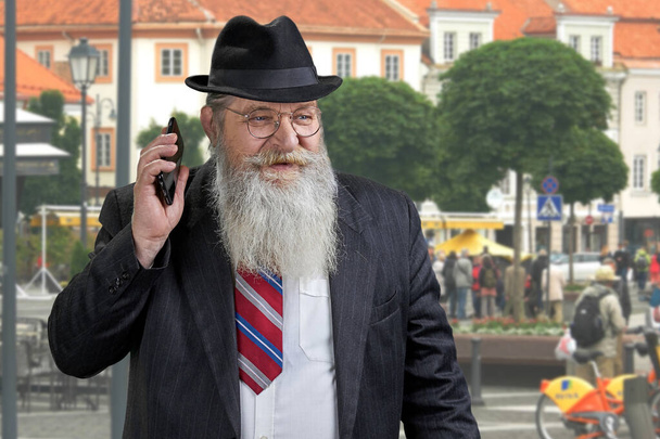 Starszy brodaty biznesmen w kapeluszu wykonuje telefon i odwraca wzrok stojąc na zewnątrz. - Zdjęcie, obraz