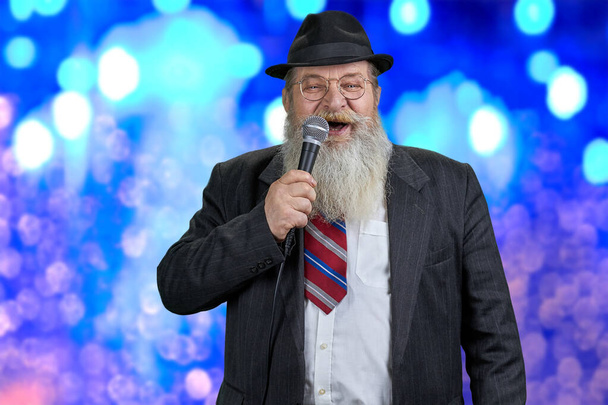 Oudere entertainer draagt pak en hoed pratend in microfoon. - Foto, afbeelding