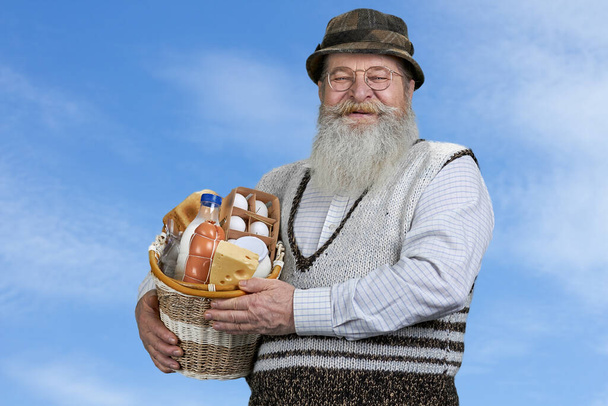 Senior man holding basket with dairy products and looking at camera on blue sky background. - Valokuva, kuva