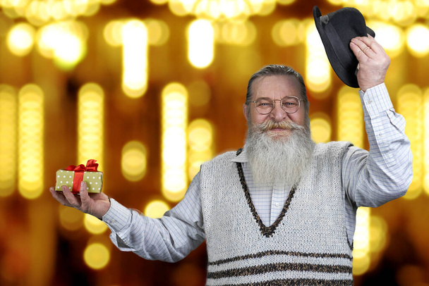 Красивый пожилой мужчина представляет подарочную коробку на фоне золотых боке огней. - Фото, изображение