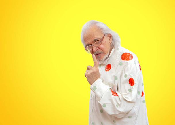 Ένας γέρος κλόουν χωρίς μακιγιάζ με φωτεινά ρούχα σε ένα φωτεινό κίτρινο φόντο δείχνει τους αντίχειρες και διδάσκει τη ζωή - Φωτογραφία, εικόνα