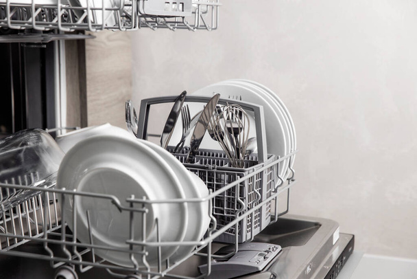 Открытая посудомоечная машина с чистой посудой внутри, столовые приборы, очки, посуда на кухне - Фото, изображение