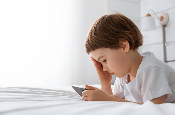 5 year baby boy lays on bed and plays with a smartphone. Close-up portrait. Holds in hands cell phone. Concept of online education. White room and clothes. Telephone user. Headache. Save child vision - Photo, image