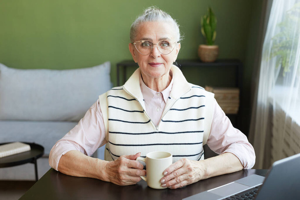 Onnellinen kypsä nainen silmälasit ja rento vaatteita ottaa teetä pöytä - Valokuva, kuva
