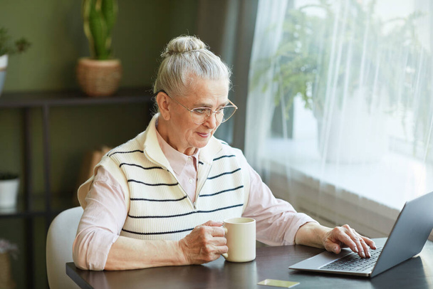 Seniorin in Freizeitkleidung trinkt Tee und blickt auf Laptop-Bildschirm - Foto, Bild