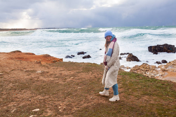 Kış denizinin kıyısında şık bir kadın yürüyor. Bej bir ceket ve şapka giymişti. Denizde fırtına vardı. - Fotoğraf, Görsel