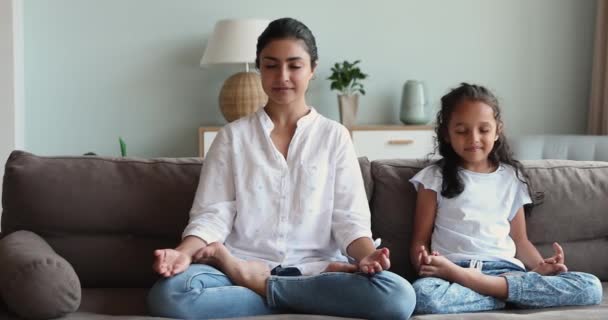 Indian mom and little daughter meditating seated on couch - Imágenes, Vídeo