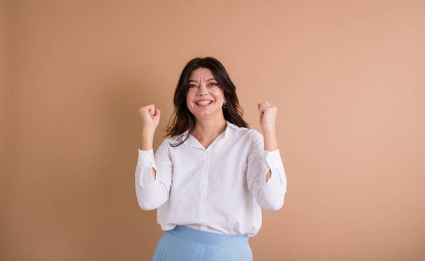 a happy brunette woman in a white shirt stands and shows success with her hands on a beige background with a place for text - Fotó, kép