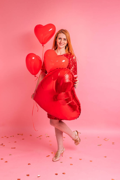 Beautiful redhead girl with red heart baloon posing. Happy Valentine's Day concept. Studio photo of beautiful ginger girl dancing on pink background. - Фото, зображення