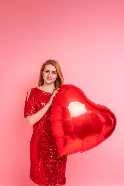 Beautiful redhead girl with red heart baloon posing. Happy Valentine's Day concept. Studio photo of beautiful ginger girl dancing on pink background. - Foto, Bild