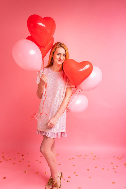 Schöne rothaarige Mädchen mit rotem Herz Luftballon posiert. Happy Valentine 's Day. Studiofoto der schönen Ingwermädchen tanzen auf rosa Hintergrund. - Foto, Bild