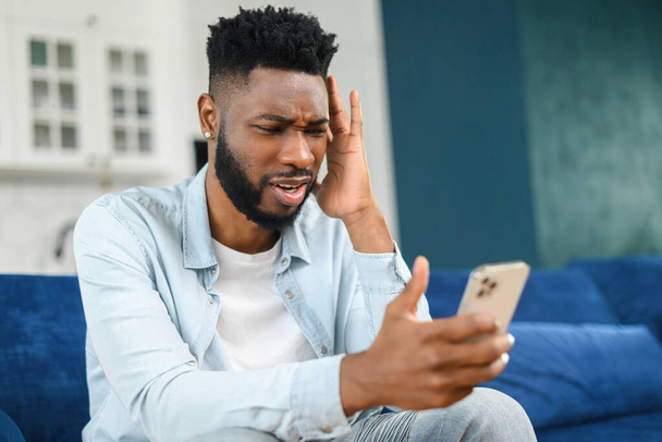 Upset multiracial businessman is talking on the smartphone. Portrait of sad failed serious male employee having conversation via video call while sitting at the sofa - Foto, Imagen