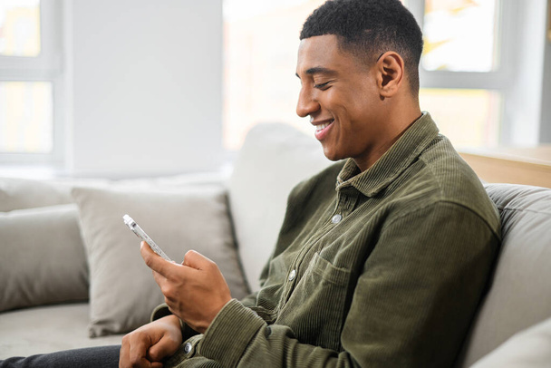 Portrait of smiling man sitting on sofa in office, using mobile phone for checking social nets, typing message for family, girlfriend, business partner - Foto, Imagen