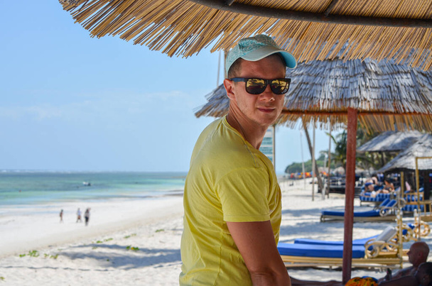 Man in a cap and sunglasses standing on the beach under an umbrella, Diani Beach, Kenya, Africa - Photo, Image