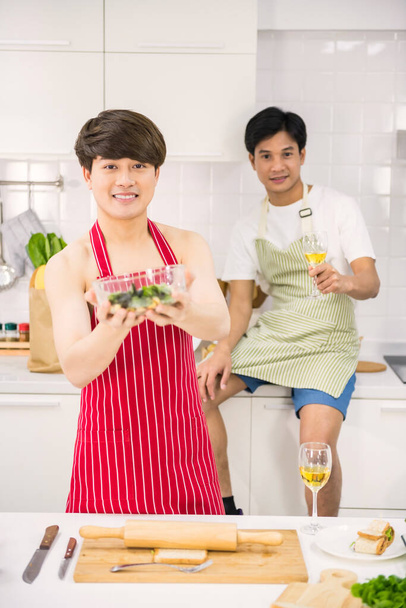 Szexi LMBT meleg fiatal ázsiai férfi piros kötényben tartsa friss salátát a kezét főzés után a konyhában, míg a partnere pirítós bor. Szép homoszexuális, azonos nemű család Valentin-napon. - Fotó, kép